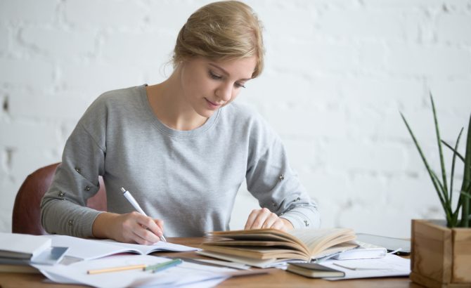 Student female performing a written task in a copybook with a pen, looking at the textbook, education concept photo, horizontal